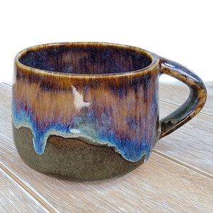 Puede incluir: Una taza de cerámica grande, marrón, azul y morada con un cuerpo ancho y redondeado y un asa. La taza tiene un efecto de glaseado de goteo.