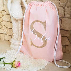 Könnte beinhalten: Ein hellrosa Kordelzugrucksack mit einem großen goldenen Glitzerbuchstaben S und dem Namen Sofia in weißer Schreibschrift. Der Rucksack verfügt über weiße geflochtene Kordelträger und besteht aus einem festen Stoff mit gerafftem Verschluss.