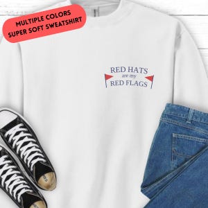 Op de afbeelding: Witte trui met de tekst "RED HATS are my RED FLAGS" in blauw en rood. De tekst wordt omringd door twee rode vlaggen met blauwe randen.