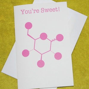 以下が含まれることがあります： 上部にピンク色の文字で「You're Sweet!」と印刷された白いグリーティングカード。テキストの下には、中央の六角形が複数のピンク色の円形ノードに接続された、化学分子構造のピンク色のグラフィックイラストが描かれています。
