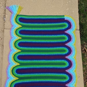 May include: A colorful crocheted rug with a rainbow pattern and fringe tassels. The rug is made with multiple shades of blue, green, purple, and yellow yarn.