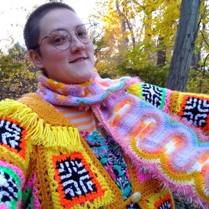 以下が含まれることがあります： カラフルな幾何学模様のクロシェ編みカーディガンと、ふわふわのピンクと白のストールを着用した人。カーディガンは、イエロー、オレンジ、ピンク、グリーン、ブラックなど、さまざまな色で作られています。ストールは波状の模様で、ふわふわの縁取りが施されています。