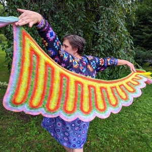 May include: A person wearing a colorful crocheted shawl with a rainbow pattern. The shawl is held up in the air, showing off its intricate design.
