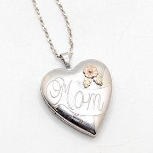 May include: Silver heart-shaped locket pendant with a delicate chain. The locket features the word "Mom" in elegant script and a small gold-toned flower accent. A thoughtful gift for Mother's Day or any special occasion.