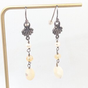 Può includere: Orecchini pendenti con un design a conchiglia. Gli orecchini presentano un ciondolo a conchiglia in metallo color argento, piccole perline rotonde e una perla ovale più grande. Gli orecchini sono esposti su un supporto color oro.