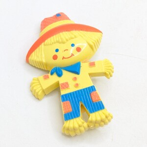 May include: A vintage, yellow, orange, and blue scarecrow figurine. The scarecrow has a yellow hat with an orange brim, a yellow body, and blue overalls. The scarecrow has a blue bow tie and rosy cheeks. The figurine is made of plastic.