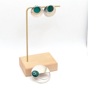 以下が含まれることがあります： 鮮やかな緑色の石がはめ込まれた円形デザインのイヤリングと、お揃いのブローチ。イヤリングは、木製の台座が付いたゴールドカラーのスタンドに展示されています。ブローチはユニークな曲線デザインです。