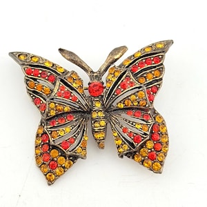 May include: A vintage butterfly brooch with a bronze-tone metal frame. The butterfly's wings are adorned with red, yellow, and orange rhinestones. A single red rhinestone accents the center of the butterfly's body. The brooch is a decorative accessory.