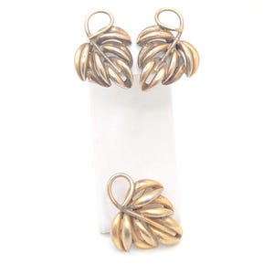 May include: A gold-toned jewelry set featuring leaf-shaped earrings and a matching pendant. The earrings are clip-on style, and the pendant has a loop for a chain. The set is displayed on a white background.