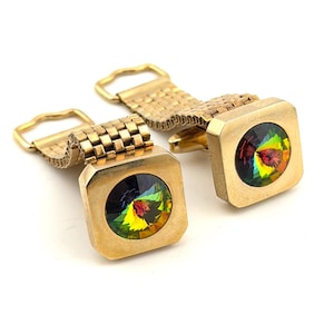May include: A pair of gold-toned cufflinks with square faces. Each cufflink features a vibrant, multi-colored, faceted gemstone. The cufflinks are connected by a gold chain and a clasp.