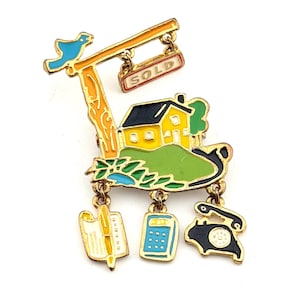 May include: A colorful enamel brooch featuring a yellow house with a black roof on a green hill. A sign reads "SOLD". Includes a blue bird, a notepad, a calculator, and a rotary phone. The brooch is gold-toned.