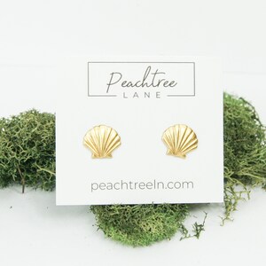 以下が含まれることがあります： 白いカードに「Peachtree Lane」と「peachtreeln.com」の文字が書かれた、金色の貝殻のスタッドピアス。