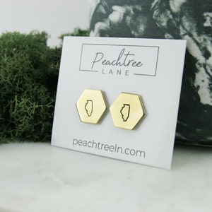 Op de afbeelding: Een paar gouden zeshoekige oorbellen met een omtrek van de staat Illinois. De oorbellen worden gepresenteerd op een witte kaart met de tekst "Peachtree Lane" en "peachtreeln.com".