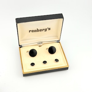 May include: A set of two round black cufflinks and three matching round black studs presented in a black and cream-colored box. The interior lid features the text renberg's and the base insert displays the brand name SWANK in black lettering.