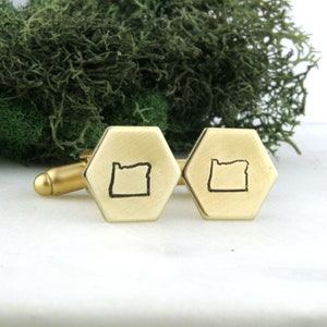 May include: Gold-coloured cufflinks with a hexagonal face. Each cufflink features a black outline of the state of Oregon. The cufflinks are set against a backdrop of green moss.