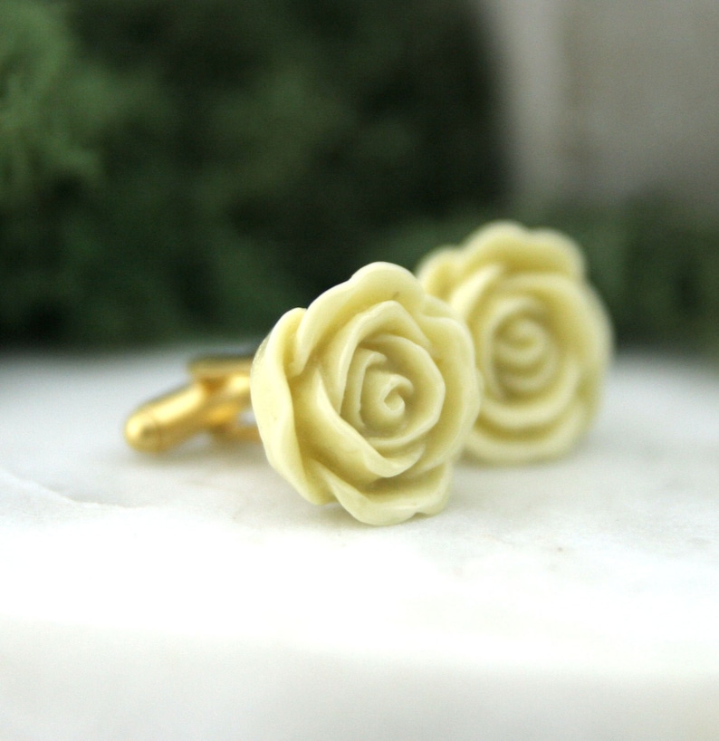 K&ouml;nnte beinhalten: Ein Paar Manschettenkn&ouml;pfe mit einem zarten Design aus elfenbeinfarbenen Rosen. Die Rosen sind mit filigranen Details gestaltet und haben eine glatte, polierte Oberfl&auml;che.