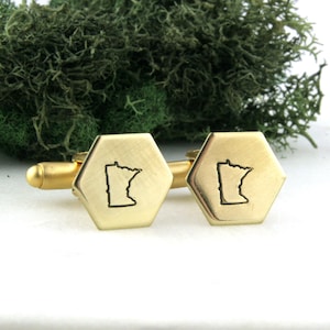 May include: A pair of gold cufflinks with a hexagonal shape. Each cufflink features an engraved outline of the state of Minnesota.