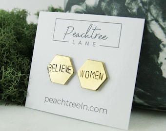 May include: A pair of gold hexagonal stud earrings with the words "BELIEVE" and "WOMEN" engraved on them. The earrings are displayed on a white card with the text "Peachtree Lane" and "peachtreeln.com".
