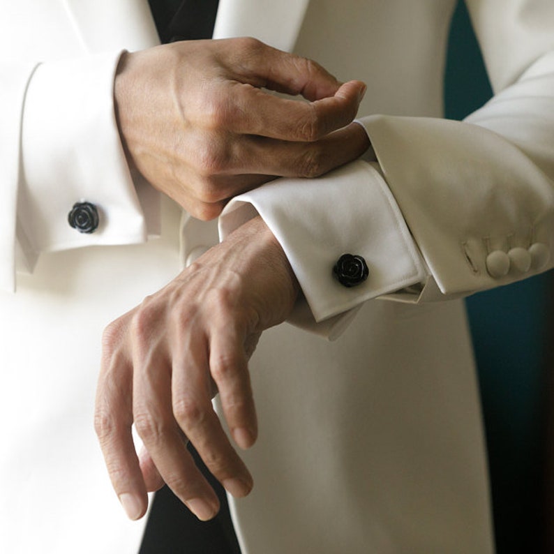 K&ouml;nnte beinhalten: Eine Person tr&auml;gt eine wei&szlig;e Smokingjacke mit schwarzen Rosenmanschettenkn&ouml;pfen. Die Person justiert die Manschettenkn&ouml;pfe an ihrem linken Handgelenk.