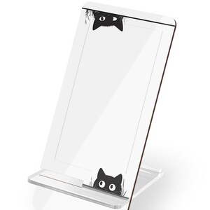 May include: White phone stand with a black cat peeking out from behind the stand. The cat has large eyes and is looking to the right. The stand is made of acrylic and has a clear base.