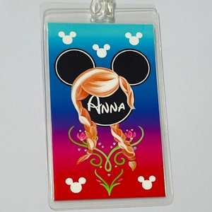 May include: A clear plastic luggage tag with a colourful design featuring a Mickey Mouse head with Anna from Frozen's hair and the name "Anna" written in white. The background is a gradient of blue, purple, and red with white Mickey Mouse ears at the top and bottom.