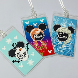 May include: Three clear plastic luggage tags with colourful Disney Frozen character designs. The tags feature Olaf, Anna, and Elsa with Mickey Mouse ears. The tags have the names "Olaf", "Anna", and "Elsa" printed on them.