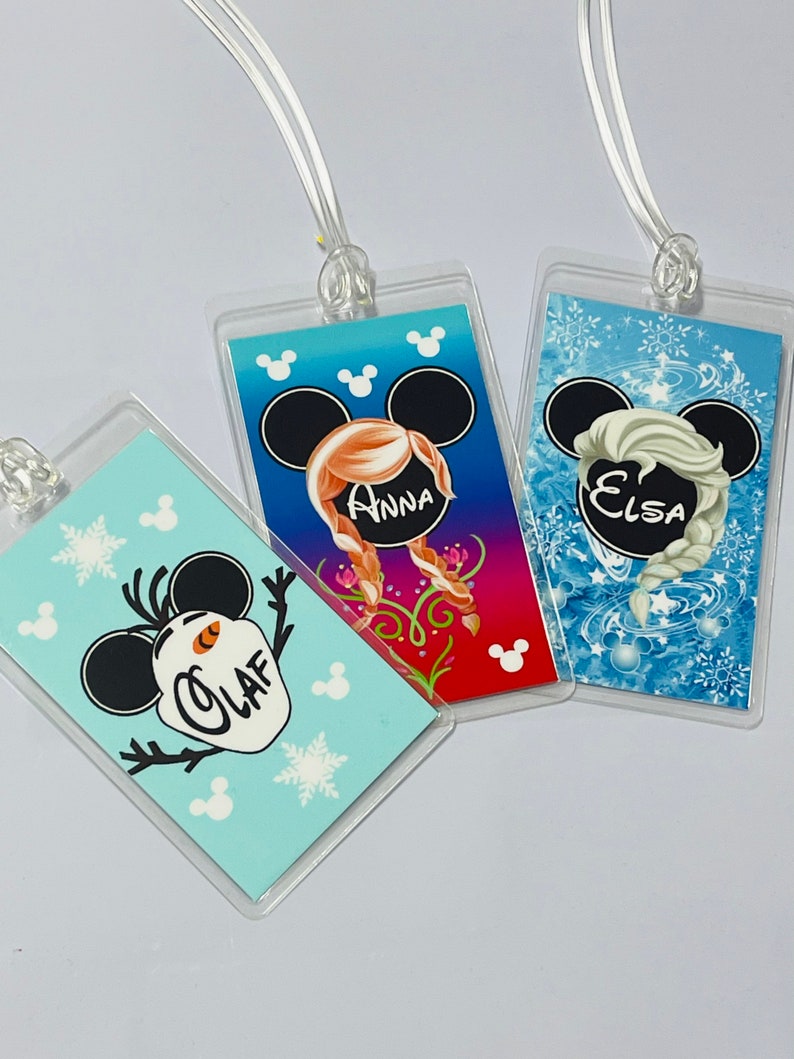 May include: Three clear plastic luggage tags with colourful Disney Frozen character designs. The tags feature Olaf, Anna, and Elsa with Mickey Mouse ears. The tags have the names "Olaf", "Anna", and "Elsa" printed on them.