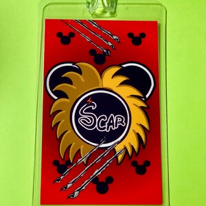 May include: A red luggage tag with a black and gold lion head design. The lion's mane is made of gold and the head is black with a white lightning bolt. The word "SCAR" is written in white letters on a black circle in the center of the lion's head. The tag is surrounded by black Mickey Mouse heads and silver claws.
