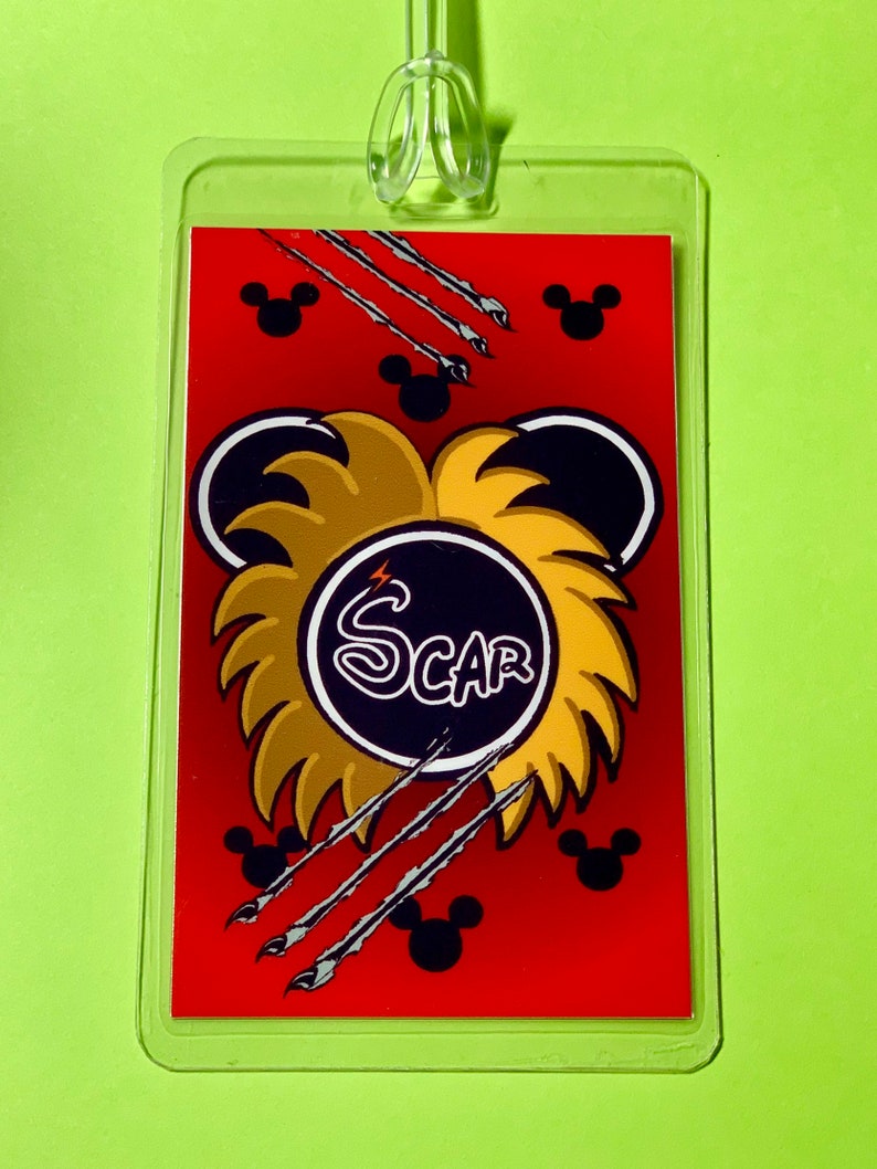May include: A red luggage tag with a black and gold lion head design. The lion's mane is made of gold and the head is black with a white lightning bolt. The word "SCAR" is written in white letters on a black circle in the center of the lion's head. The tag is surrounded by black Mickey Mouse heads and silver claws.