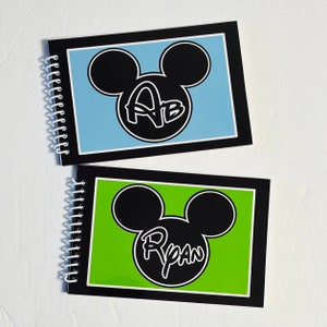 Op de afbeelding: Twee zwarte spiraalnotitieblokken met Mickey Mouse-oren en een gepersonaliseerde naam. Het bovenste notitieblok heeft een lichtblauwe achtergrond met de naam "Ab" in zwart. Het onderste notitieblok heeft een groene achtergrond met de naam "Ryan" in zwart.