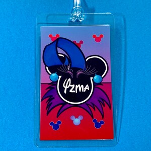 May include: A purple and red Mickey Mouse themed luggage tag with the name "Azma" written in white. The tag has blue and purple accents and features Mickey Mouse ears.