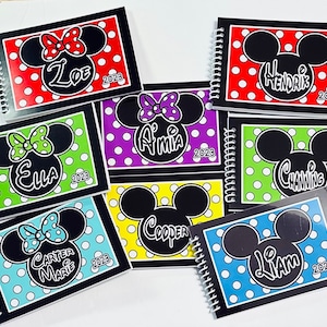 Op de afbeelding: Zeven zwarte spiraalgebonden notitieboekjes met kleurrijke stippenachtergronden en een zwart-wit Mickey Mouse of Minnie Mouse hoofd op elke omslag. De namen Ella, Carter Marie, Amia, Cooper, Liam, Channing en Zoe staan ​​op de omslagen samen met het jaar 2023.