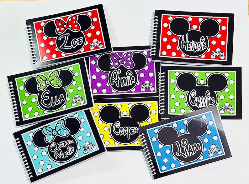 Op de afbeelding: Zeven zwarte spiraalgebonden notitieboekjes met kleurrijke stippenachtergronden en een zwart-wit Mickey Mouse of Minnie Mouse hoofd op elke omslag. De namen Ella, Carter Marie, Amia, Cooper, Liam, Channing en Zoe staan ​​op de omslagen samen met het jaar 2023.