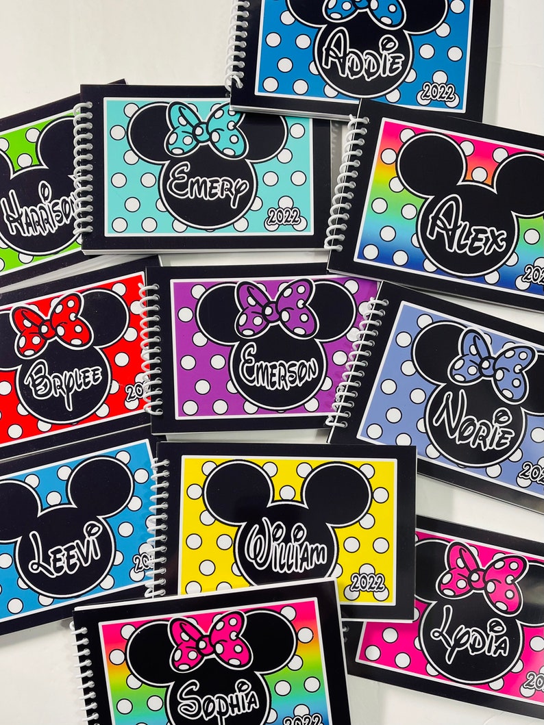 Op de afbeelding: Een verzameling kleurrijke, gepersonaliseerde notitieboekjes met een Mickey Mouse-silhouet met stippen en een strik. Elk notitieboekje heeft een andere naam en het jaar 2022 staat op de omslag.