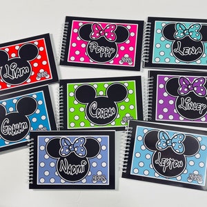 Pode incluir: Oito cadernos negros com espiral e capas de bolinhas coloridas e orelhas do Mickey Mouse. Cada caderno tem um nome diferente na capa, incluindo Liam, Poppy, Lena, Corban, Linley, Graham, Naomi e Lepton. O ano de 2022 est&aacute; impresso em cada caderno.