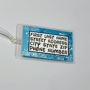 May include: A clear plastic luggage tag with a blue and white snowflake pattern. The tag has black text that reads "FIRST LAST NAME STREET ADDRESS CITY STATE ZIP PHONE NUMBER".