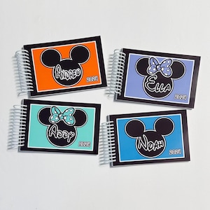 Op de afbeelding: Vier gepersonaliseerde spiraalnotitieblokken met Mickey Mouse en Minnie Mouse oren op de voorkant. De notitieblokken zijn oranje, blauw, paars en turquoise. De namen "Andrea", "Ella", "Addy" en "Noah" staan op de voorkant van elk notitieblok, samen met het jaar "2024".