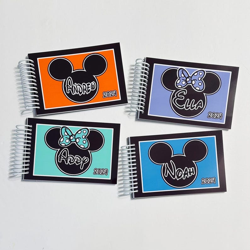 Op de afbeelding: Vier gepersonaliseerde spiraalnotitieblokken met Mickey Mouse en Minnie Mouse oren op de voorkant. De notitieblokken zijn oranje, blauw, paars en turquoise. De namen "Andrea", "Ella", "Addy" en "Noah" staan op de voorkant van elk notitieblok, samen met het jaar "2024".