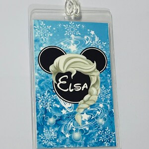 May include: A blue and white luggage tag featuring a black Mickey Mouse head silhouette. The head has a white braid with the name "Elsa" written in black inside a black circle.