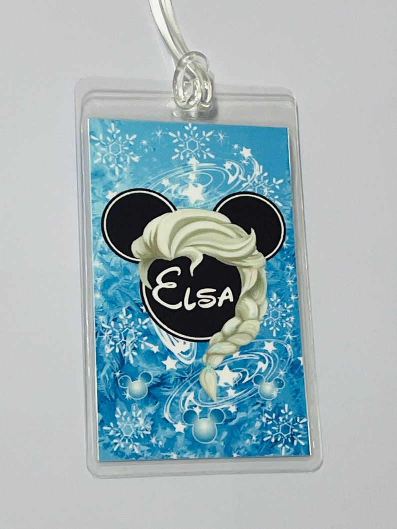 May include: A blue and white luggage tag featuring a black Mickey Mouse head silhouette. The head has a white braid with the name "Elsa" written in black inside a black circle.