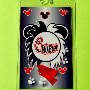 May include: A black and white luggage tag with a cartoon image of Cruella de Vil's hair, red gloves, and paw prints. The tag has a silver background with black Mickey Mouse ears and red Mickey Mouse ears.