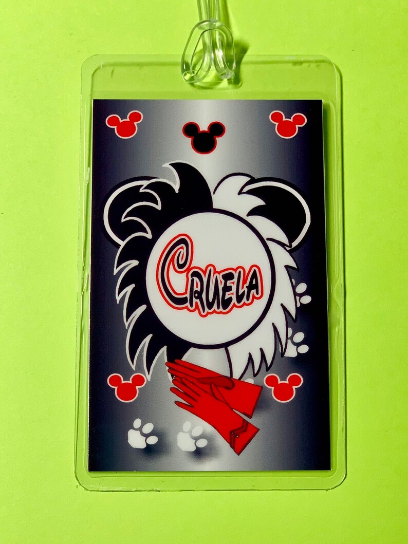 May include: A black and white luggage tag with a cartoon image of Cruella de Vil's hair, red gloves, and paw prints. The tag has a silver background with black Mickey Mouse ears and red Mickey Mouse ears.