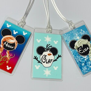May include: Three luggage tags featuring Disney's Frozen characters. The tags are clear plastic with a colourful image of Anna, Elsa, and Olaf. Each tag has a black Mickey Mouse ear design with the character's name printed in white.