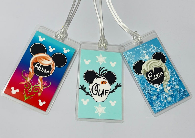 May include: Three luggage tags featuring Disney's Frozen characters. The tags are clear plastic with a colourful image of Anna, Elsa, and Olaf. Each tag has a black Mickey Mouse ear design with the character's name printed in white.