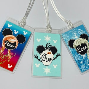 May include: Three luggage tags featuring Disney's Frozen characters. The tags are clear plastic with a colourful image of Anna, Olaf, and Elsa. Each tag has the character's name printed on it.