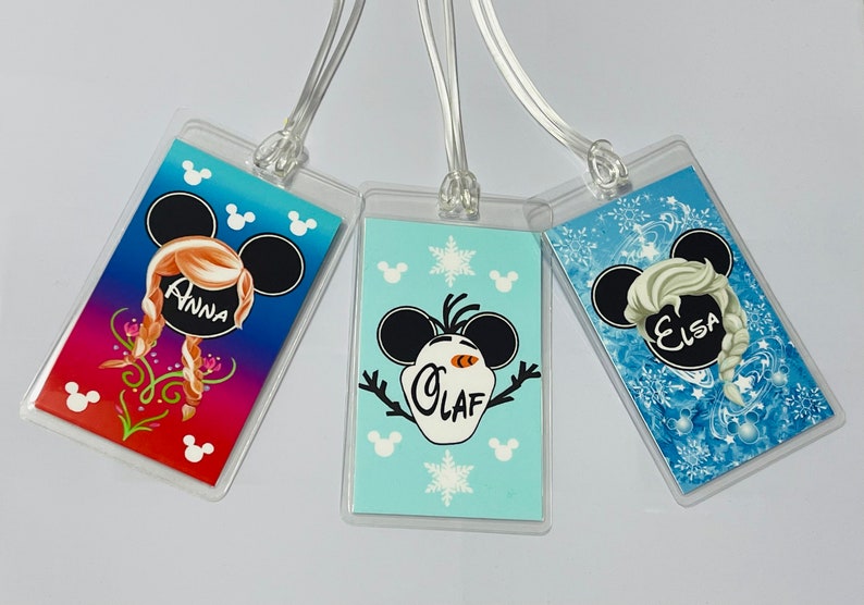 May include: Three luggage tags featuring Disney's Frozen characters. The tags are clear plastic with a colourful image of Anna, Olaf, and Elsa. Each tag has the character's name printed on it.