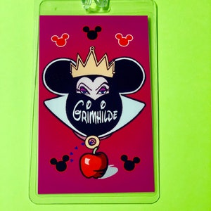 May include: A red and black Disney-themed luggage tag featuring a cartoon image of the Evil Queen from Snow White with a crown, a black mask, and a red apple. The text "Grimhilde" is written on the mask. The background is a pink color with black Mickey Mouse ears.