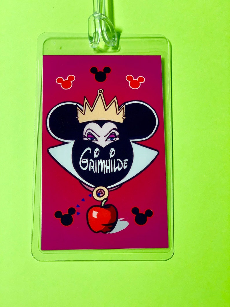 May include: A red and black Disney-themed luggage tag featuring a cartoon image of the Evil Queen from Snow White with a crown, a black mask, and a red apple. The text "Grimhilde" is written on the mask. The background is a pink color with black Mickey Mouse ears.