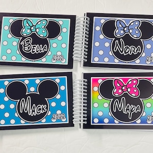Op de afbeelding: Vier zwarte spiraalgebonden notitieboekjes met een Mickey Mouse of Minnie Mouse ontwerp. Elk notitieboekje heeft een andere achtergrondkleur en een andere naam op de voorkant: Bella, Nora, Mack en Mapa. Het jaar 2022 is op elk notitieboekje gedrukt.