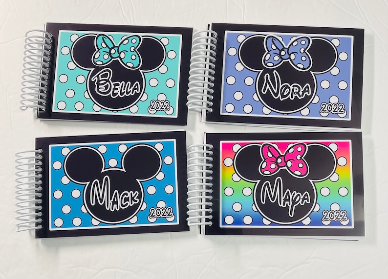 Op de afbeelding: Vier zwarte spiraalgebonden notitieboekjes met een Mickey Mouse of Minnie Mouse ontwerp. Elk notitieboekje heeft een andere achtergrondkleur en een andere naam op de voorkant: Bella, Nora, Mack en Mapa. Het jaar 2022 is op elk notitieboekje gedrukt.